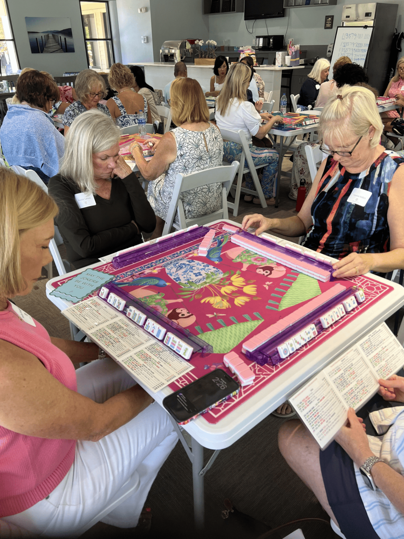 A busy room of players seated around pink and purple mahjong tables